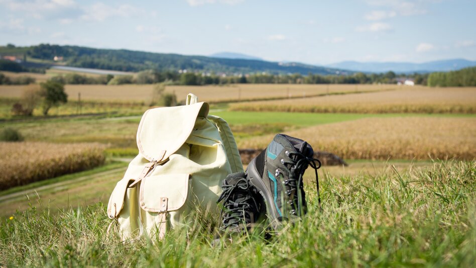 Wanderschuhe und Wanderrucksack liegen in der Wiese mit Blick auf die steirische Hügellandschaft