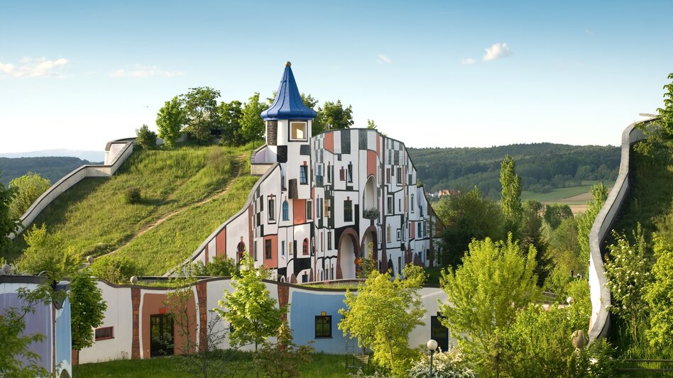 Verspieltes, farbenfrohes Gebäude der Therme Rogner Bad Blumau im unverkennbaren Stil von Friedensreich Hundertwasser, eingebettet in die grüne Hügellandschaft der Oststeiermark. | © Rogner Bad Blumau, Hundertwasser Architekturprojekt