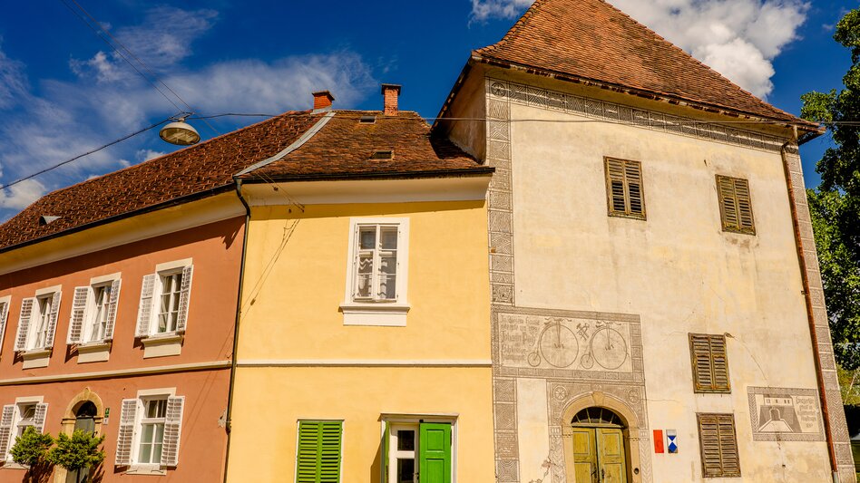 Historische Lehrstätte Puch in Bad Radkersburg mit hellen Fassaden, grünen Fensterläden, steilen Ziegeldächern und einem hohen Gebäudeteil mit Sonnenuhr.