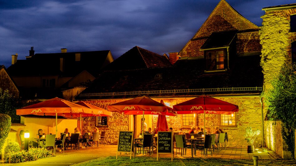 Stimmungsvoller Gastgarten in Bad Radkersburg mit roten Sonnenschirmen und Außengastronomie vor historischer Architektur am Abend.