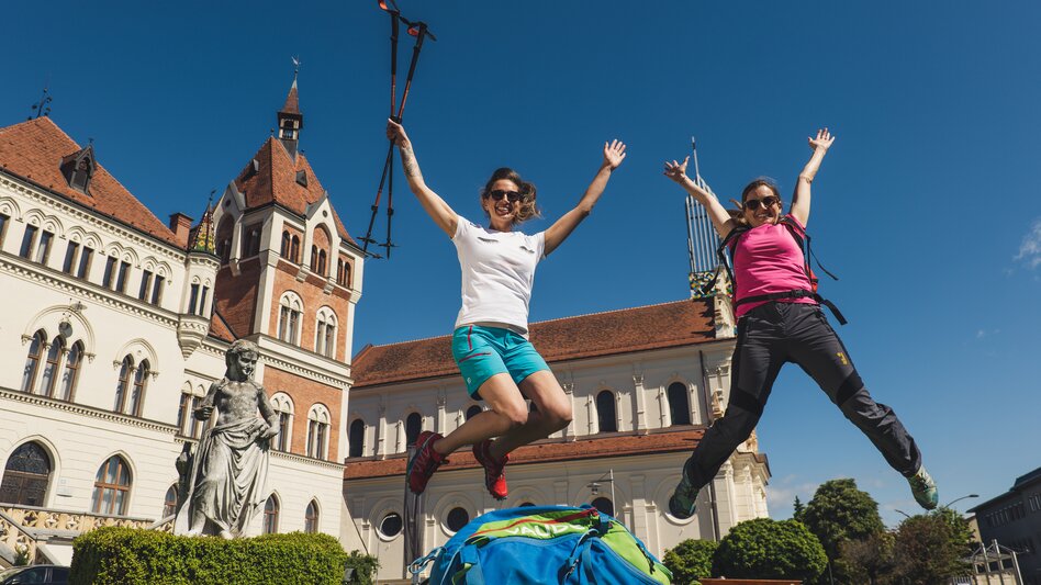 Freunde springen für dem Feldbacher Rathaus und Kirchturm durch die Luft | © Tourismusverband Thermen- & Vulkanland I Bernhard Bergmann