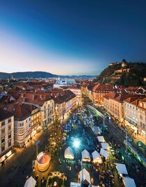 Panoramablick über den festlich beleuchteten Adventmarkt am Grazer Hauptplatz mit Weihnachtsbaum, Verkaufsständen und Blick auf den Uhrturm – ein pulsierendes Zentrum winterlicher Magie.