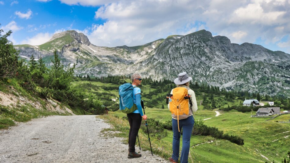 Pärchen von hinten auf Schotterweg Richtung Almhütten unterwegs, im Hintergrund eine Bergkulisse