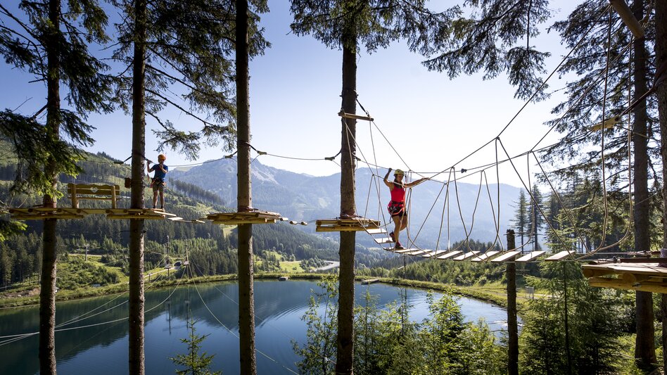 Zwei Personen klettern auf dem Adventure Park vor einem See in den Bäumen