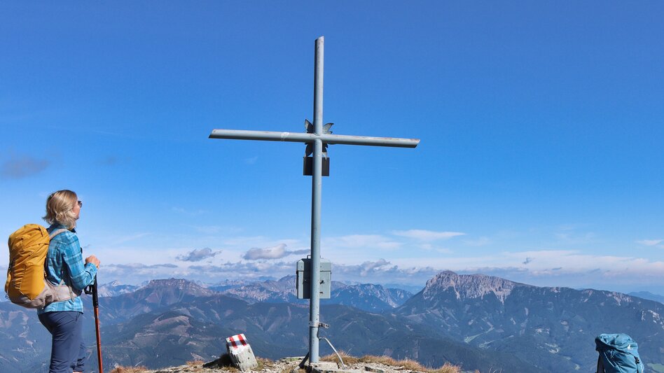 Frau steht vor Gipfelkreuz und schaut es an, dahinter die schöne Aussicht auf die umliegenden Berge | © WEGES OG