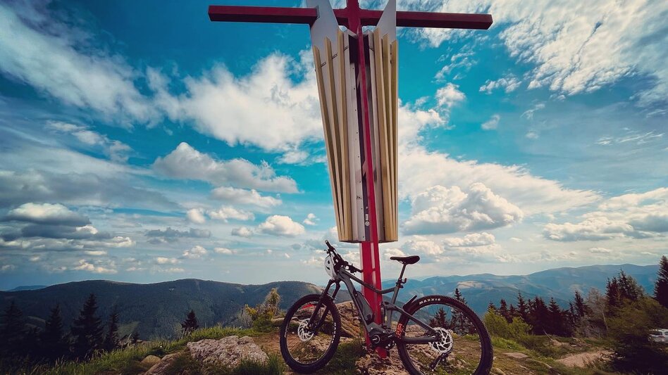 Das Mugelgipfelkreuz, davor steht ein Fahrrad mit einer herrlichen Aussicht auf die gegenüber liegenden Berge