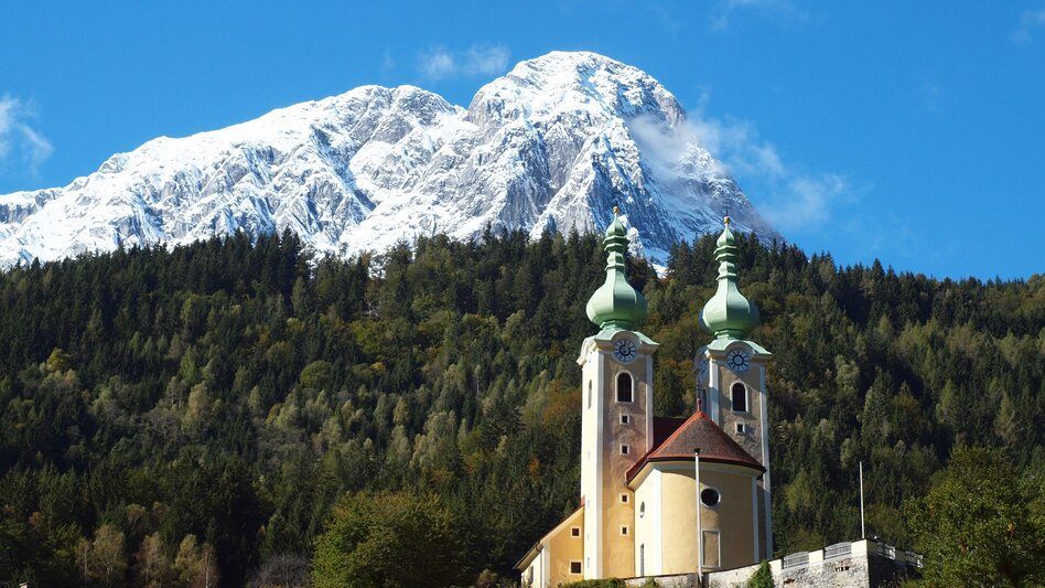 Eine Kirche umgeben vom Wald, dahinter ein mit schneebedeckter Berg