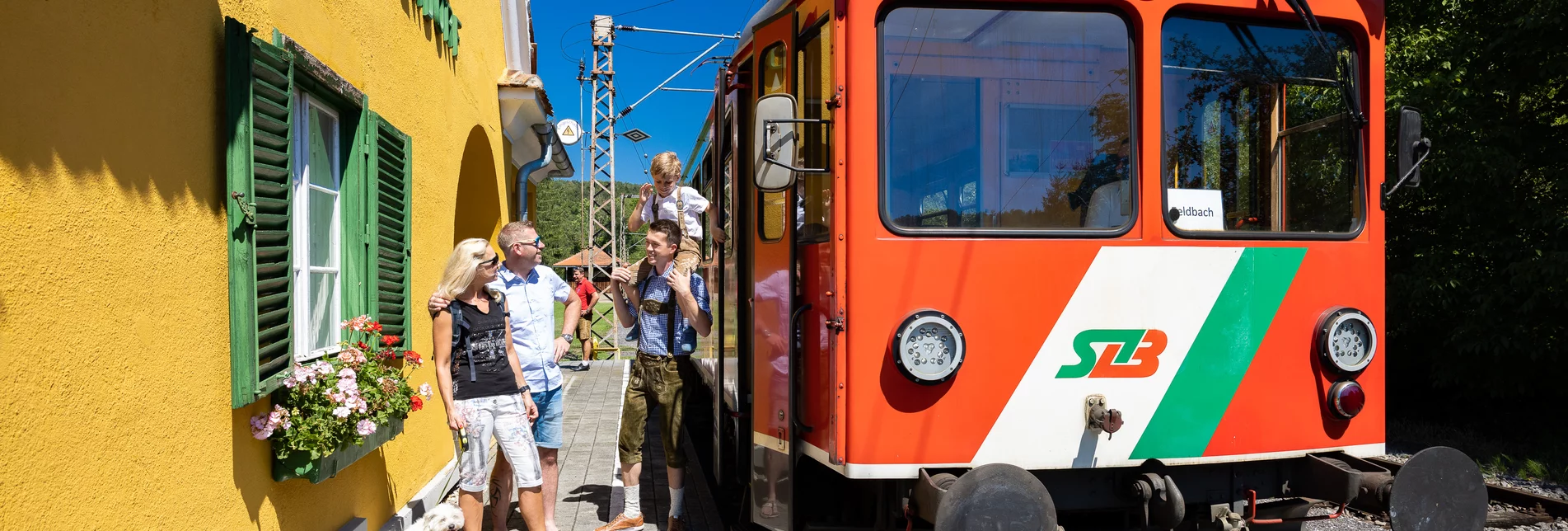 Es ist ein sonniger Tag, der Himmel ist wolkenlos. Vor dem roten Zug der Steiermarkbahn, der vor dem Bahnhaus Halt macht, unterhalten sich drei Personen. | © StB