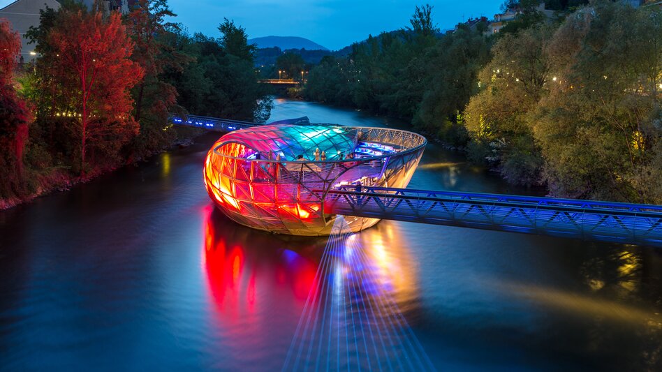Die bunt beleuchtete Murinsel in Graz bei Nacht, eingebettet in den Fluss Mur, mit umliegenden Bäumen und zwei Fußgängerbrücken. | © Graz Tourismus