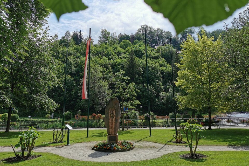 <p>Der gepflegte Kurpark in Bad Aussee mit grünen Bäumen und bunten Blumen. Im Mittelpunkt steht der Mittelpunktstein, daneben steht die österreichische Flagge.</ | © TVB Ausseerland - Salzkammergut, www.ausseerland.at