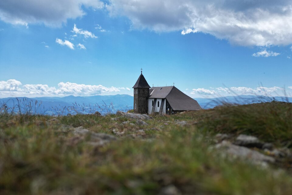Eine kleine Kirche steht auf einer Wiese mit Blick auf die Berge. Der Himmel ist blau und leicht bewölkt. | © Isabella Painhapp