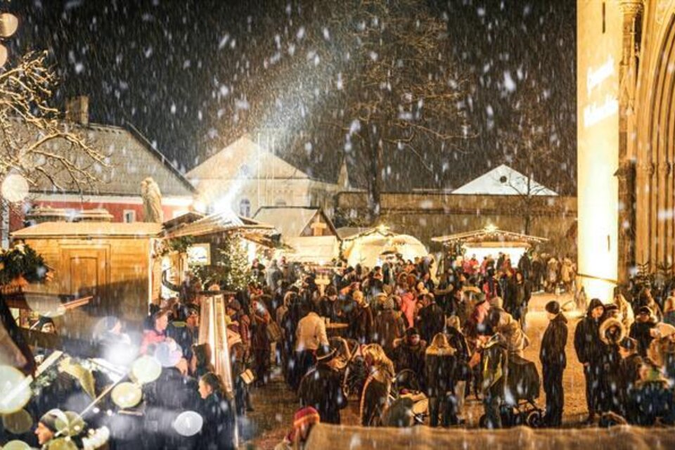 Ein festlicher Weihnachtsmarkt bei Nacht, beleuchtet mit Lichtern und umgeben von Schnee. Menschen genießen die festliche Atmosphäre und die Stände mit verschiedenen Angeboten.