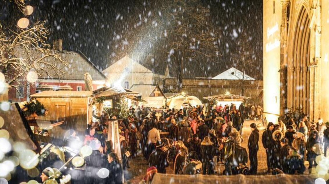 Ein festlicher Weihnachtsmarkt bei Nacht, beleuchtet mit Lichtern und umgeben von Schnee. Menschen genießen die festliche Atmosphäre und die Stände mit verschiedenen Angeboten.