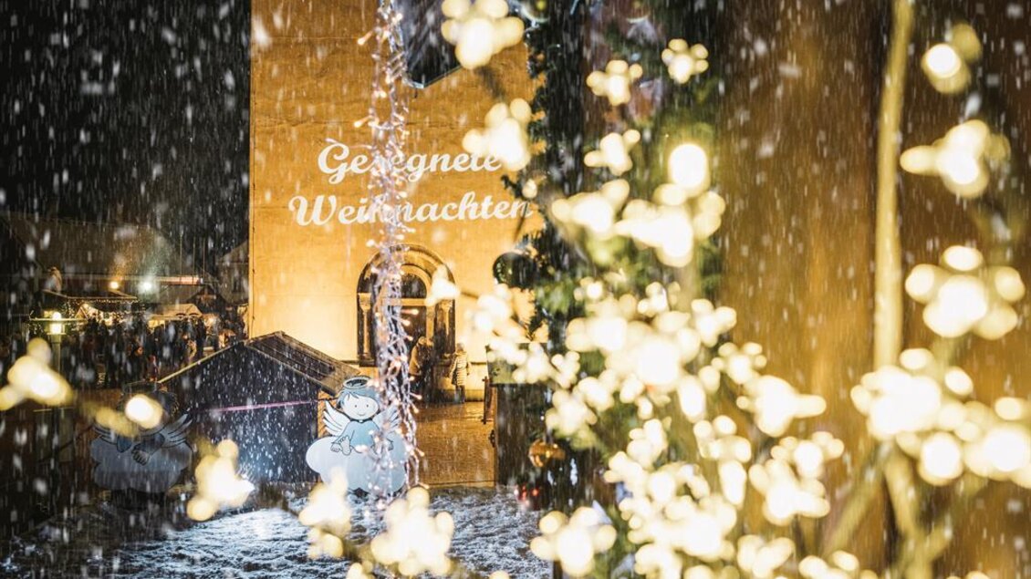 Eine festliche Winterlandschaft mit schneebedeckten Straßen und geschmückten Lichtern. Im Hintergrund ist ein Gebäude zu sehen, das mit "Gesegnete Weihnachten" beleuchtet ist.