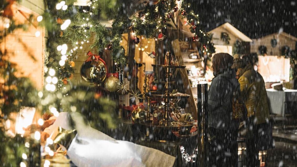 Ein festlicher Weihnachtsmarkt mit hellen Lichtern und dekorierten Ständen. Es schneit, während Menschen die Stände besuchen und Geschenke betrachten.