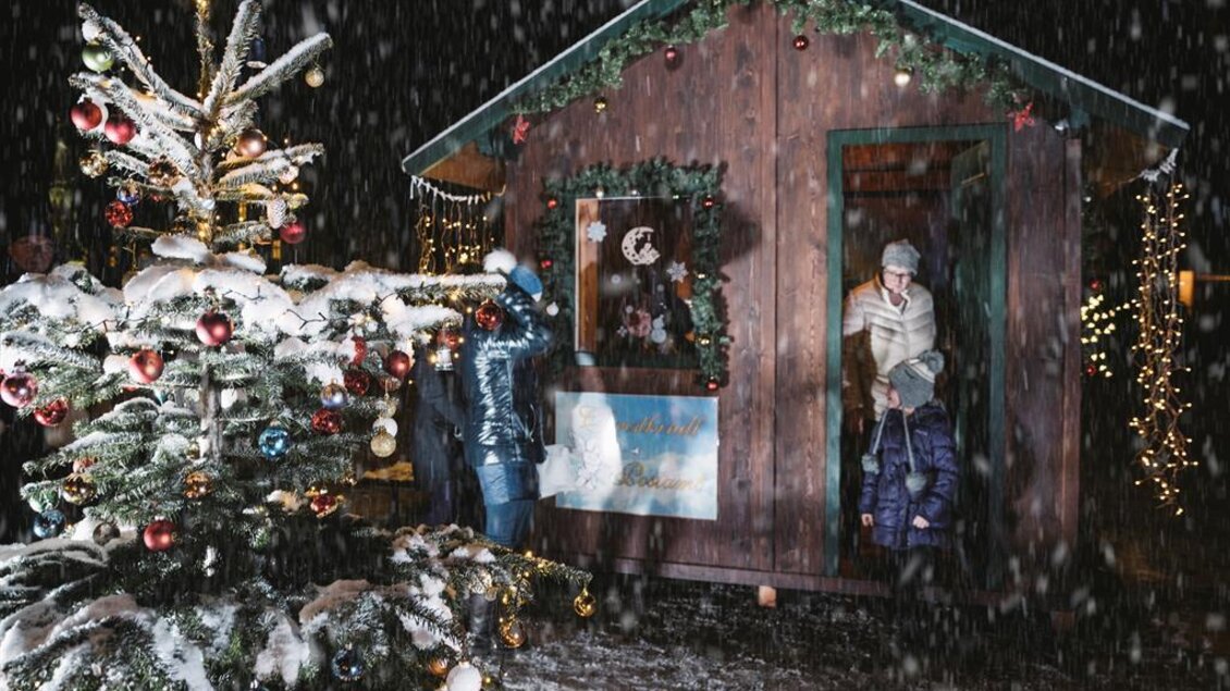 Eine festlich dekorierte Holzlaube steht vor einem geschmückten Weihnachtsbaum. Es schneit und die Umgebung ist mit Lichtern erleuchtet.