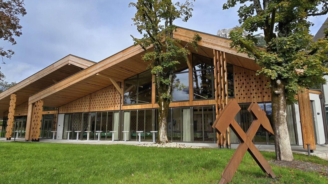 Der moderne Veranstaltungssaal im Herzen von Altaussee aus viel Holz und Glas. Im Vordergrund ist der grüne Kurpark der Gemeinde am Altausseer See zu sehen, der von einer Skulptur mit dem Bergmannszeichen aus Cortenstahl in seiner charakteristischen rostbraunen Farbe geschmückt wird, welche am rechten Bildrand zu sehen ist | © Stephanie Bor