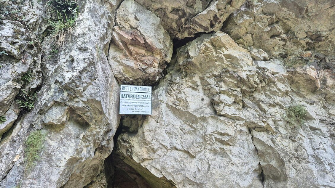 Ein großer Felsen mit einer Höhle und einem Schild darüber. Der Eingang zur Höhle ist von steilen Felswänden umgeben. | © Andrea Stelzer