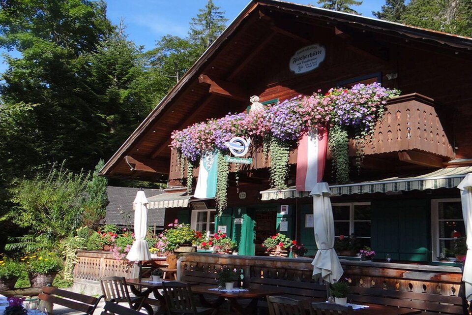 Ein charmantes, rustikales Gasthaus umgeben von Bäumen und bunten Blumen. Der Balkon ist mit Pflanzen dekoriert, und Tische stehen im Freien zum Verweilen bereit. | © Fischerhütte/Syen