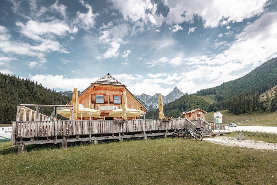 Eine gemütliche Berghütte mit einer Terrasse und Blick auf die umliegenden Berge. Im Vordergrund sind Fahrräder und eine grüne Wiese zu sehen. | © Thomas Sattler