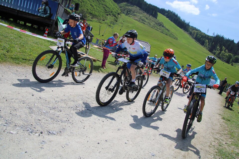 Eine Gruppe von Kindern fährt begeistert mit dem Mountainbike auf einem Schotterweg. Im Hintergrund sind grüne Hügel und Zuschauer zu sehen. | © Johann Schröcker