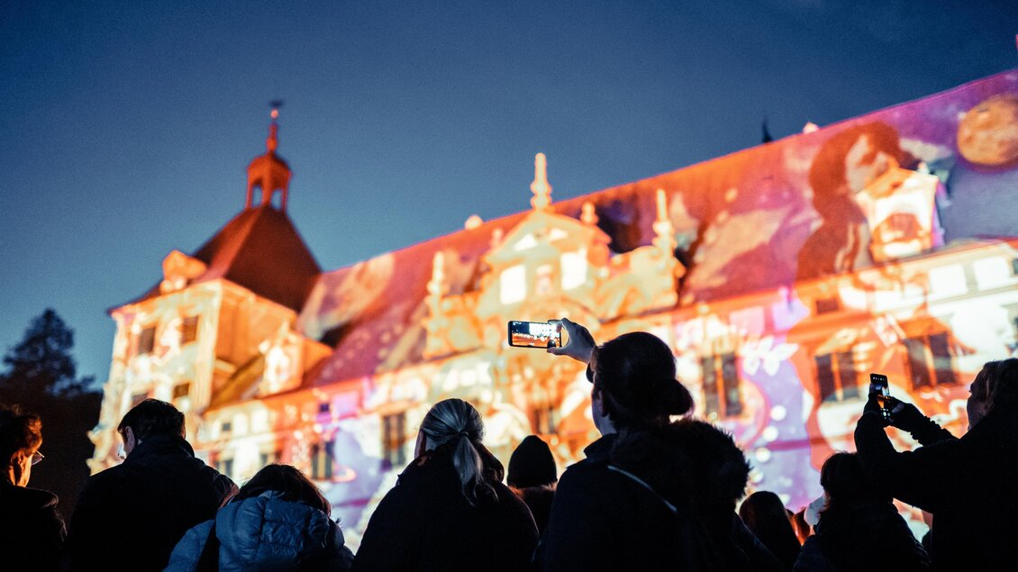 Eine Gruppe von Menschen steht vor einem Gebäude, das mit bunten Lichtern projiziert wird. Es ist Abend und die Stimmung ist festlich. | © (c) Alex Koch