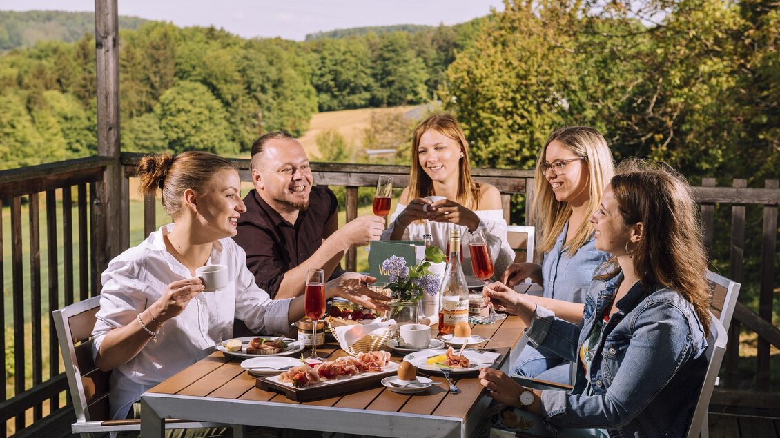 Eine Gruppe von fünf Personen sitzt an einem Tisch im Freien und genießt ein Essen. Es gibt verschiedene Speisen und Getränke, während sie miteinander anstoßen und die Natur genießen. | © Bernhard Bergmann