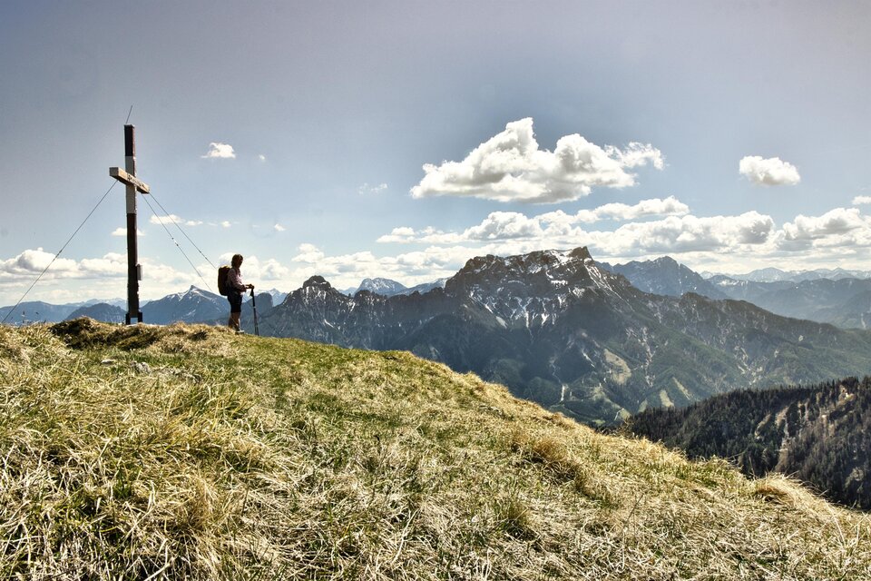 Eine Person steht an einem Gipfelkreuz und schaut auf eine beeindruckende Berglandschaft. Der Himmel ist klar mit wenigen Wolken.