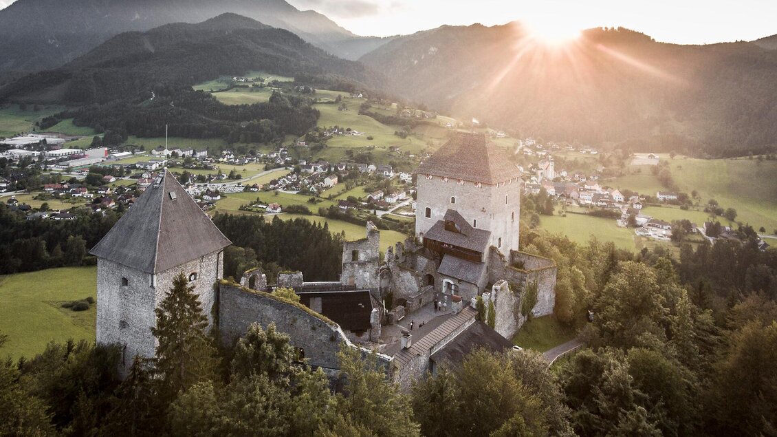 Eine alte Burgruine liegt eingebettet in eine grüne Landschaft. Die Sonne geht hinter den Bergen auf und beleuchtet die historischen Mauern. | © Stefan Leitner