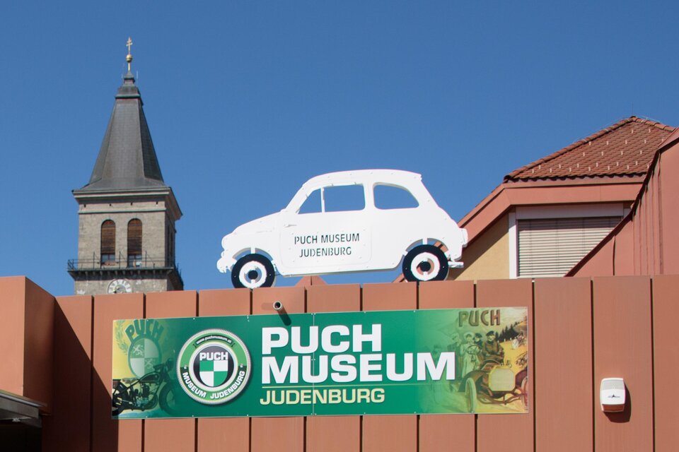 Vier historische Autos sind ausgestellt, angeordnet auf einem bunten Banner. Die Fahrzeuge sind in verschiedenen Farben und zeigen die zeitlose Schönheit der Automobilgeschichte. | © Foto Mitteregger