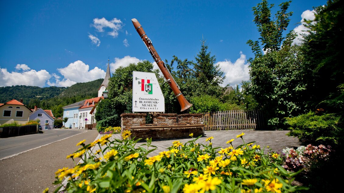 Ein fröhlicher Ortseingang mit einer großen Holzfigur und bunten Blumen im Vordergrund. Der Himmel ist blau mit einigen Wolken. | © Holzwelt Murau