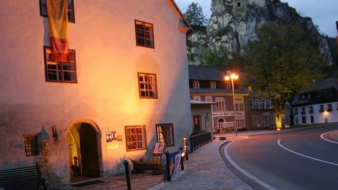 Eine historische Straße mit einem beleuchteten Gebäude und einer Burg im Hintergrund. Die Umgebung ist abends mit sanftem Licht und Natur umgeben. | © Marktgemeinde Thörl