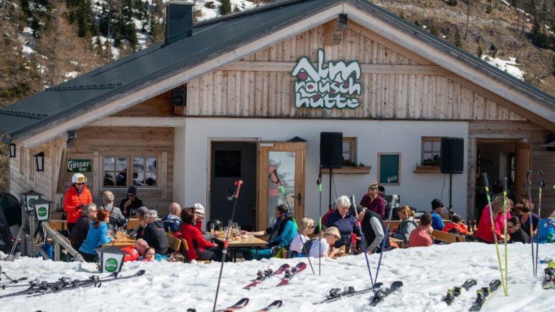 Eine gemütliche Skihütte in den Bergen, umgeben von Schnee. Viele Gäste genießen ihre Zeit draußen mit Essen und Getränken. | © Paller