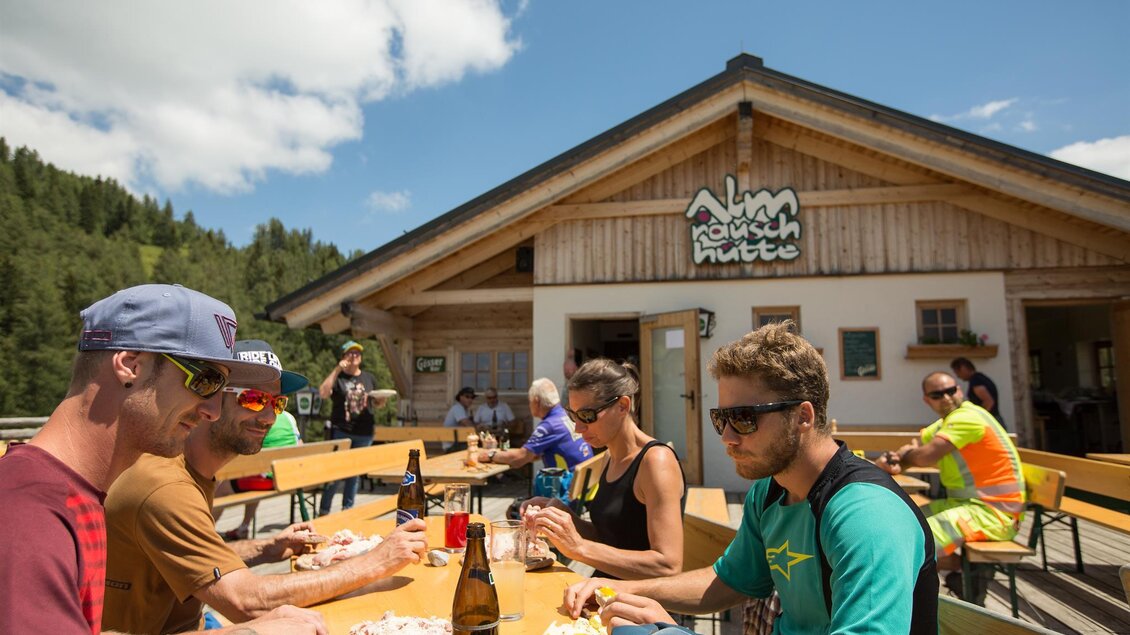 Eine Gruppe von Freunden sitzt an einem Tisch im Freien und genießt ein Essen. Im Hintergrund steht ein gemütliches Bergrestaurant umgeben von grüner Natur. | © Klaus Morgenstern