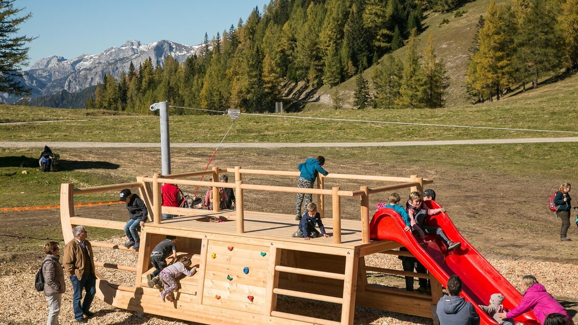 Ein Spielplatz mit einem großen Holzspielgerät und einer Rutsche. Kinder spielen und klettern im Freien, umgeben von Bergen und Bäumen. | © Klaus Morgenstern