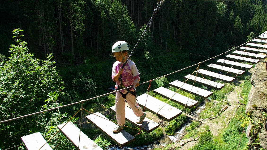 Ein Kind geht vorsichtig über einen Hängeweg aus Holzplanken, umgeben von Bäumen und einer Schlucht. Es trägt einen Helm und ist mit einem Sicherheitsgurt gesichert. | © Ewald Walder