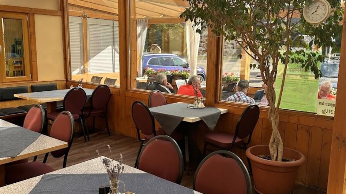 Ein gemütliches Restaurant mit Holzmöbeln und großen Fenstern. Pflanzen und gedeckte Tische schaffen eine einladende Atmosphäre.