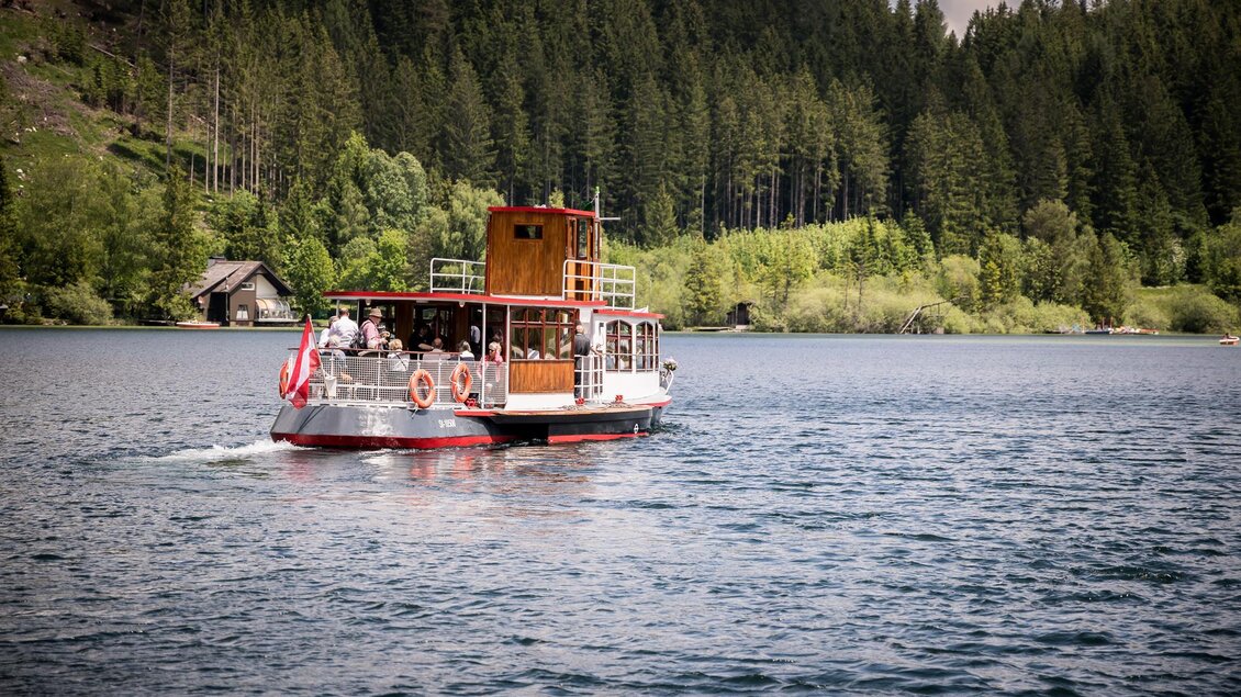 Ein Boot fährt über einen ruhigen See, umgeben von üppigen, grünen Wäldern. Am Ufer sind Bäume und eine kleine Hütte zu sehen. | © TV Hochsteiermark