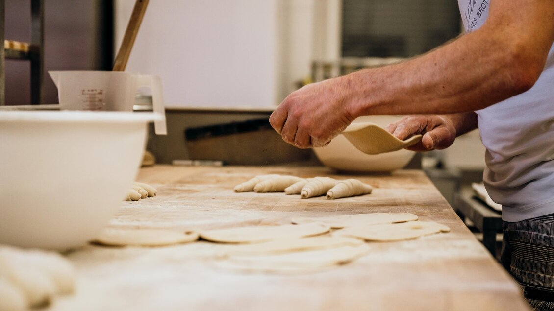 Ein Bäcker arbeitet an einem Tisch, auf dem Teiglinge liegen. In der Nähe steht eine große Schüssel und eine Holzlatte. | © GenussReich