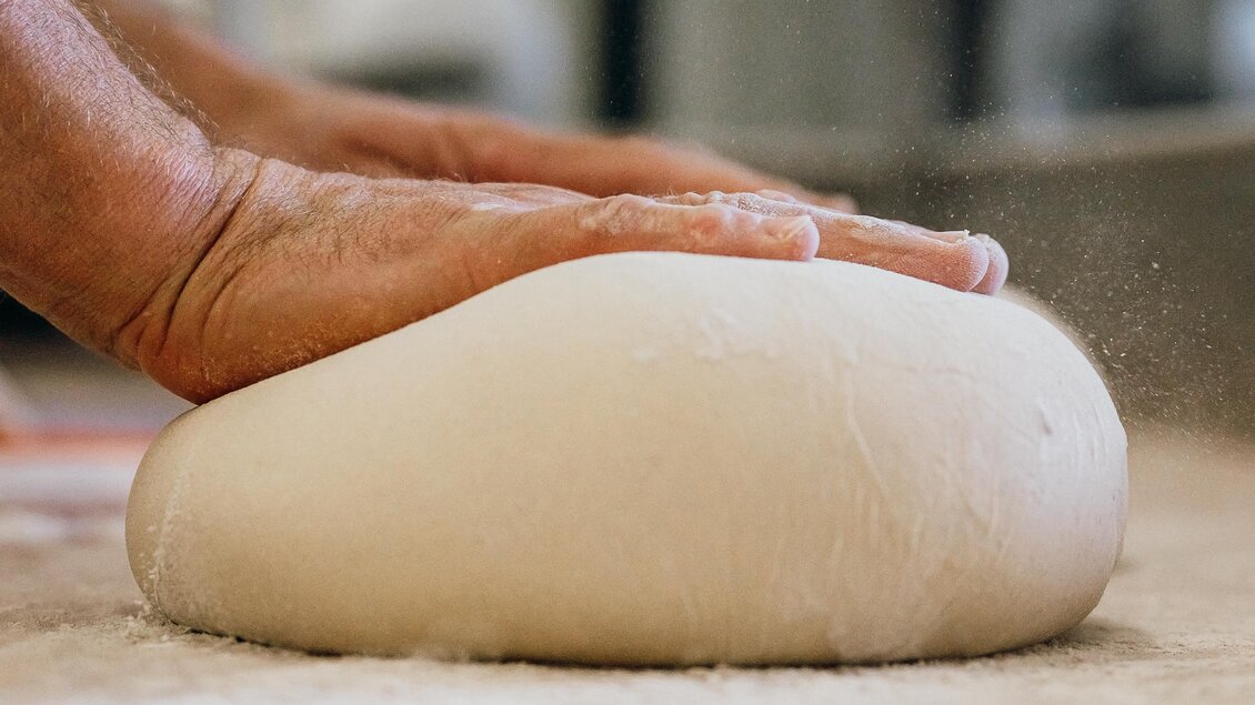 Ein Handwerker knetet Teig auf einer bemehlten Arbeitsfläche. Der Teig hat eine weiche, runde Form. | © GenussReich
