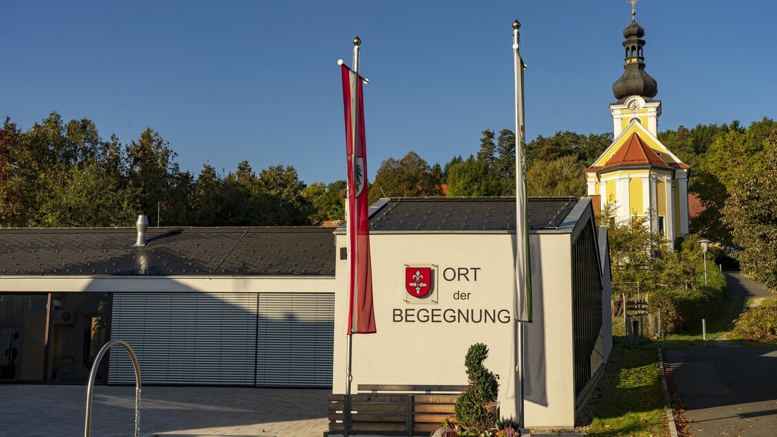 Ein modernes Gebäude mit der Beschriftung "Ort der Begegnung" ist im Vordergrund. Im Hintergrund steht eine kirchenartige Struktur mit einer auffälligen Kuppel. | © Kurkommission Bad Blumau