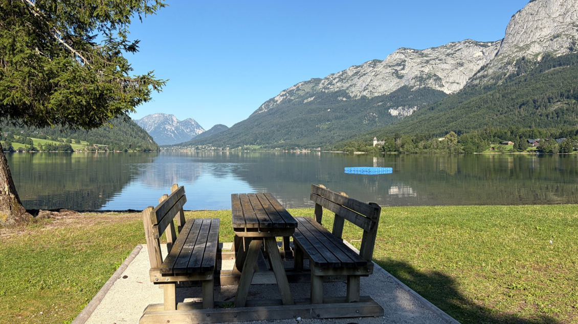 <p>Zwei Holzbänke mit einem Tisch direkt am Ufer des Grundlsees in Gößl. Im Hintergrund sind die Berge des toten Gebirges und ein blauer Himmel zu sehen.</p> | © Tourismusverband Ausseerland