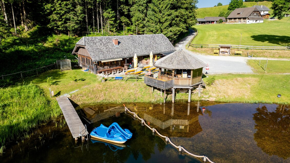 Eine idyllische Naturansicht mit einer Hütte am Wasser und umliegenden Bäumen. Ein Tretboot liegt am Ufer und die Landschaft ist grün und einladend. | © TV Murau