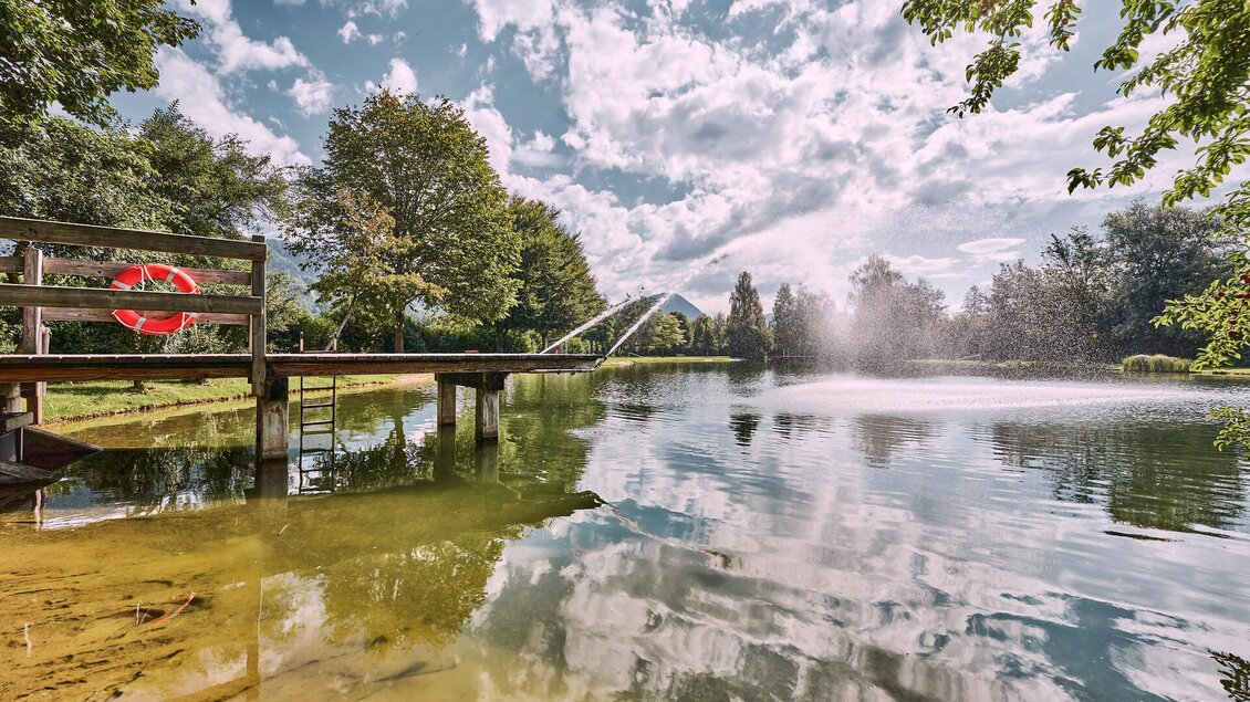 Ein ruhiger See umgeben von grünen Bäumen und sanften Hügeln. Der Himmel ist klar mit einigen weißen Wolken. | © Thomas Sattler