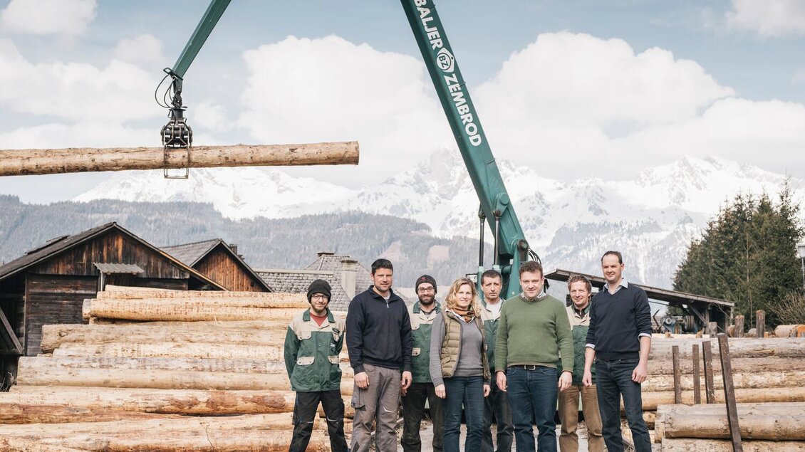 Eine Gruppe von sieben Personen steht vor einem Holzlager mit Baumstämmen. Im Hintergrund sind Berge und ein Kran sichtbar, der Holz hebt. | © Stefan Leitner