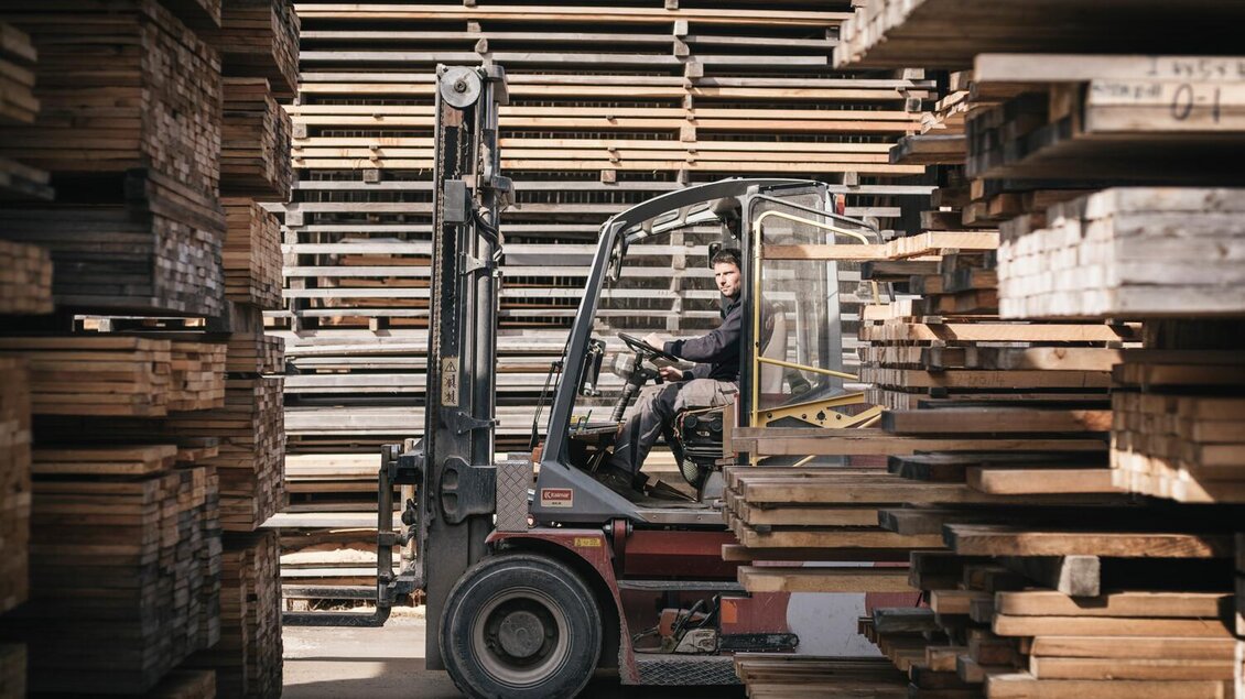 Ein Gabelstapler bewegt sich zwischen Holzstapeln in einem Lager. Im Führungssitz sitzt ein Mann, der das Fahrzeug steuert. | © Stefan Leitner