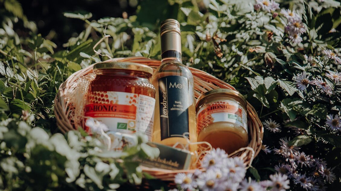 Ein Korb mit verschiedenen Lebensmitteln, darunter Marmelade und Honig, steht im Grünen. Die Produkte sind umgeben von bunten Blumen. | © GenussReich