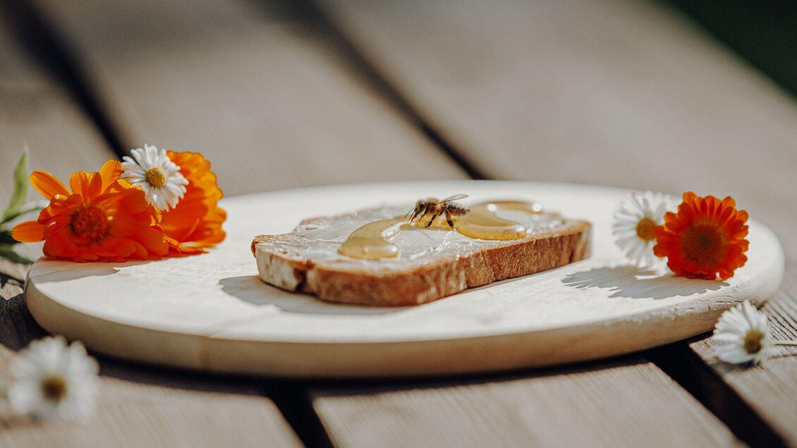 Ein Stück Brot mit Honig liegt auf einer Holzplatte. Daneben sind frische Blumen in orange und weiß arrangiert. | © GenussReich