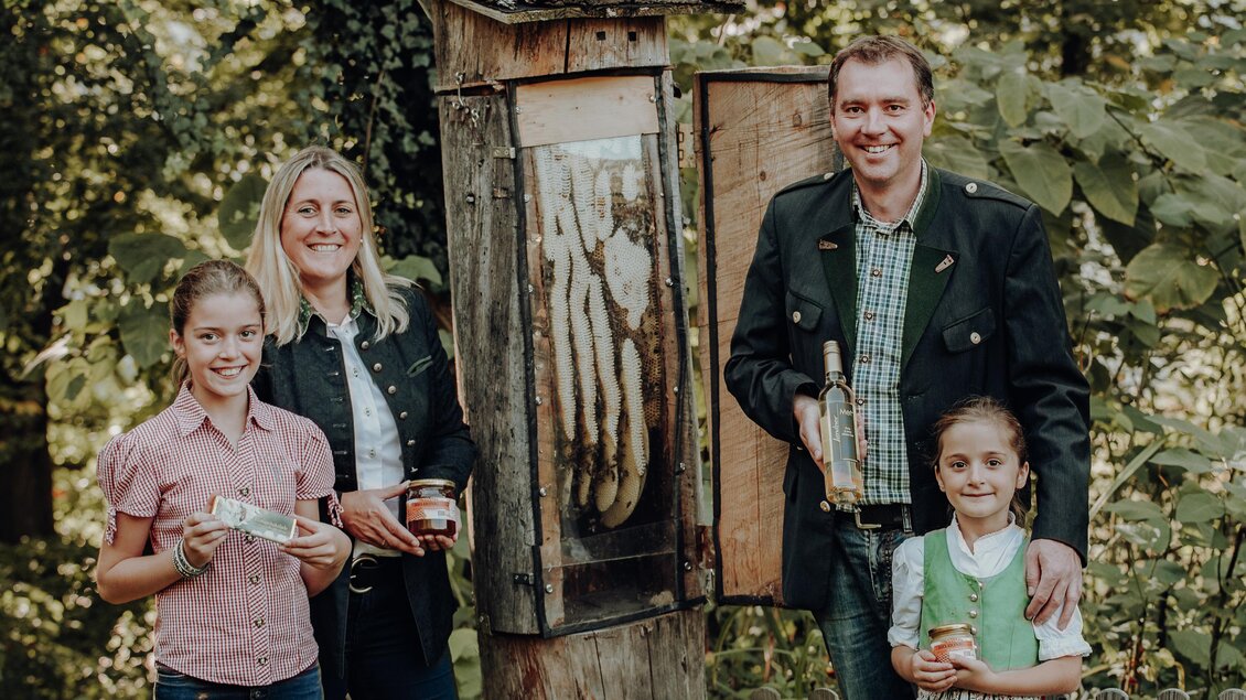 Eine Familie steht im Wald neben einem Bienenstock. Sie halten Honiggläser in der Hand und lächeln in die Kamera. | © GenussReich