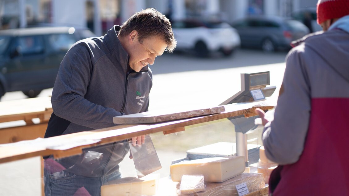 Ein Verkäufer steht an einem Marktstand und bietet Waren an. Im Hintergrund sind Autos und andere Marktbesucher zu sehen. | © Tourismusverband Murau